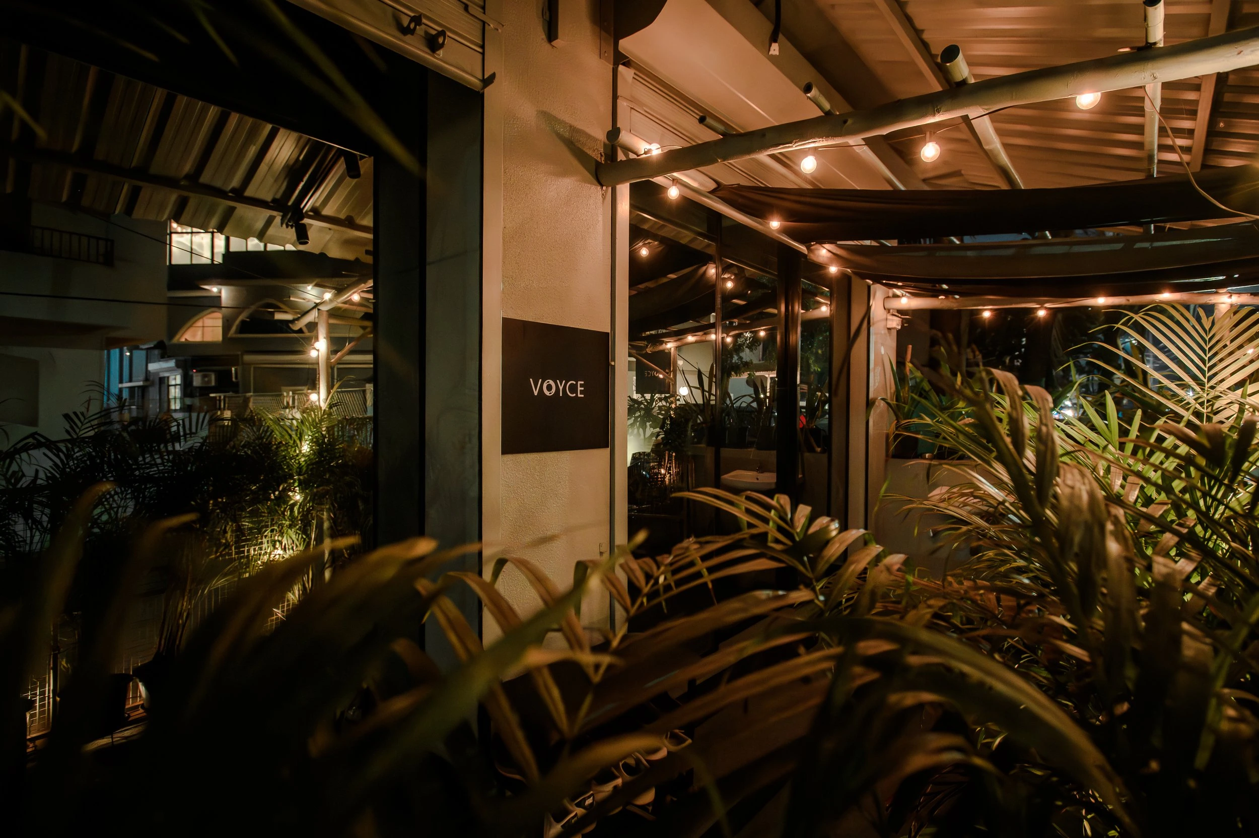 Nighttime view of a cozy outdoor patio area of a restaurant or bar named VOYCE, with warm string lights, plants, and a glass door entrance reflected in the nearby window.